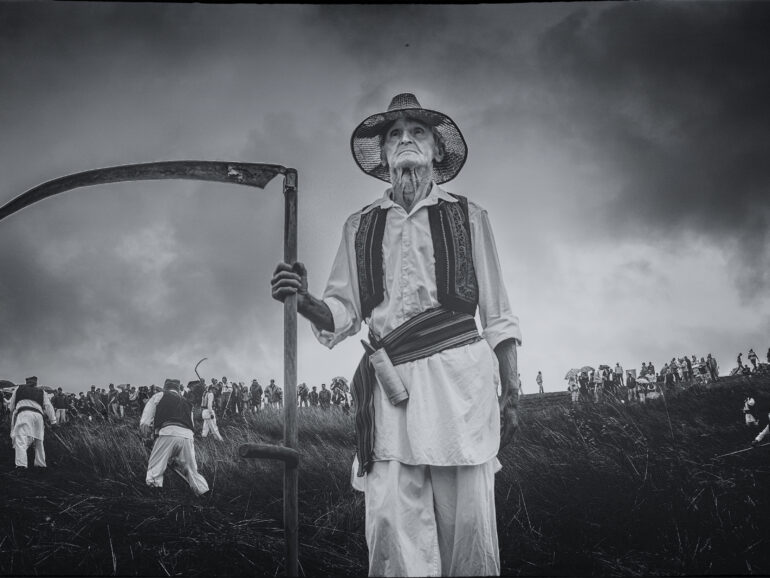 Portrait of an elderly man holding a scythe during a traditional rural gathering, with people working and observing in the background.
